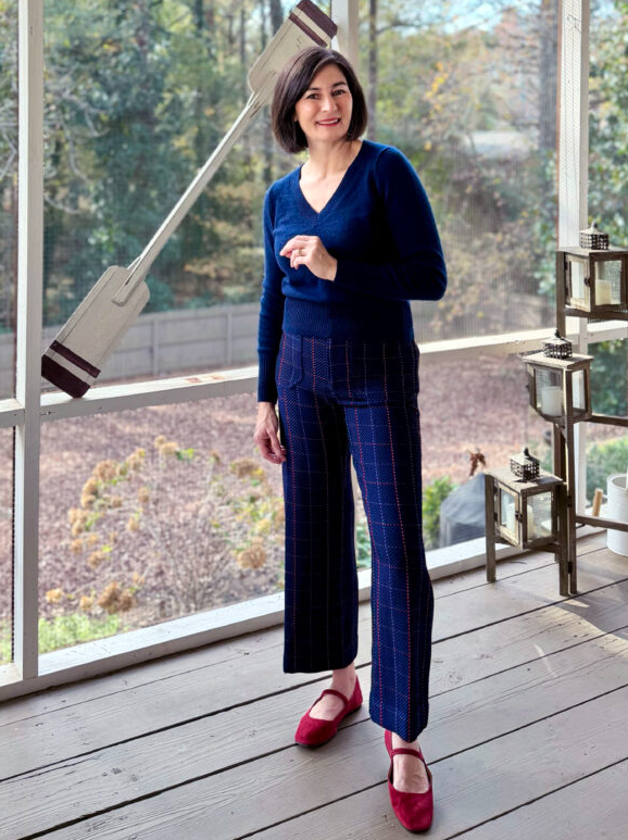 Woman indoors wearing a navy V-neck sweater with navy plaid cropped trousers, paired with red suede Mary Jane flats for a polished, comfortable look.