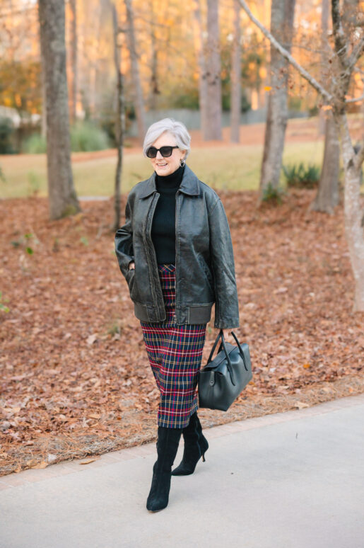 Woman wearing a black leather jacket over a black turtleneck, styled with a red-and-navy plaid midi skirt, knee-high black heeled boots, and a structured black handbag while walking outdoors in autumn.