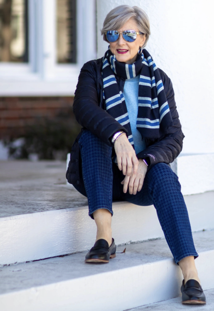 Woman wearing a black quilted jacket over a blue sweater with a navy-and-white striped scarf, patterned blue ankle pants, and black loafers, seated on a white porch step wearing sunglasses.