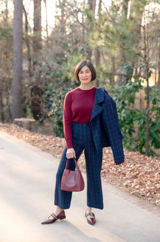 Woman wearing a burgundy knit top styled with navy plaid cropped trousers and a matching blazer draped over her shoulder, finished with burgundy leather loafers and a structured handbag while standing on a tree-lined path.