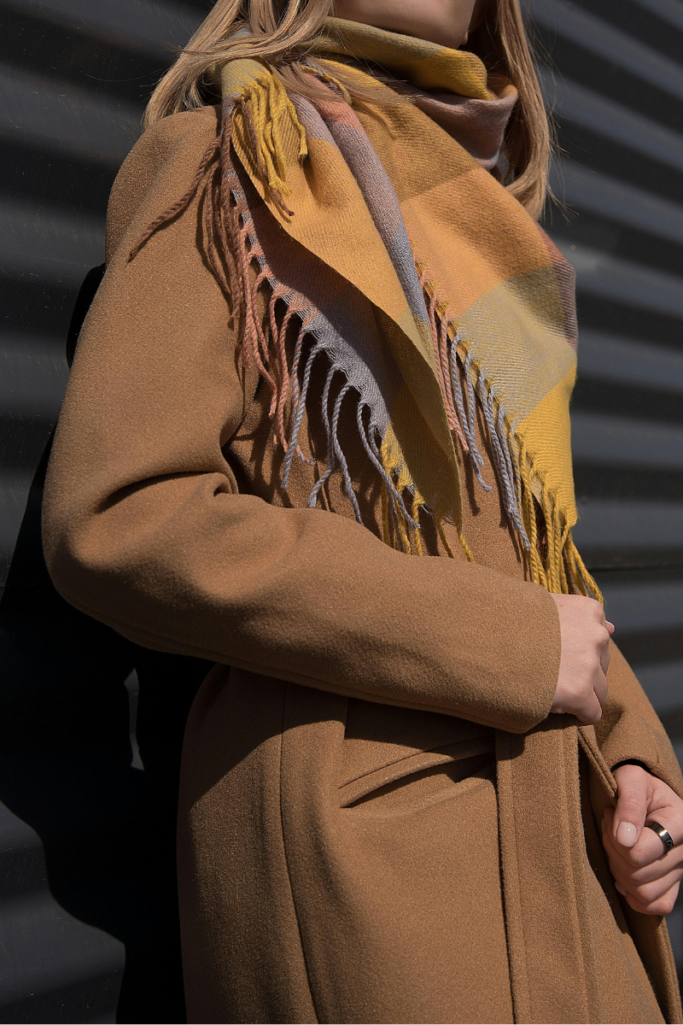Close-up of a camel wool coat styled with a large plaid scarf in warm mustard, brown, and gray tones, showing layered winter textures and fringe detail.