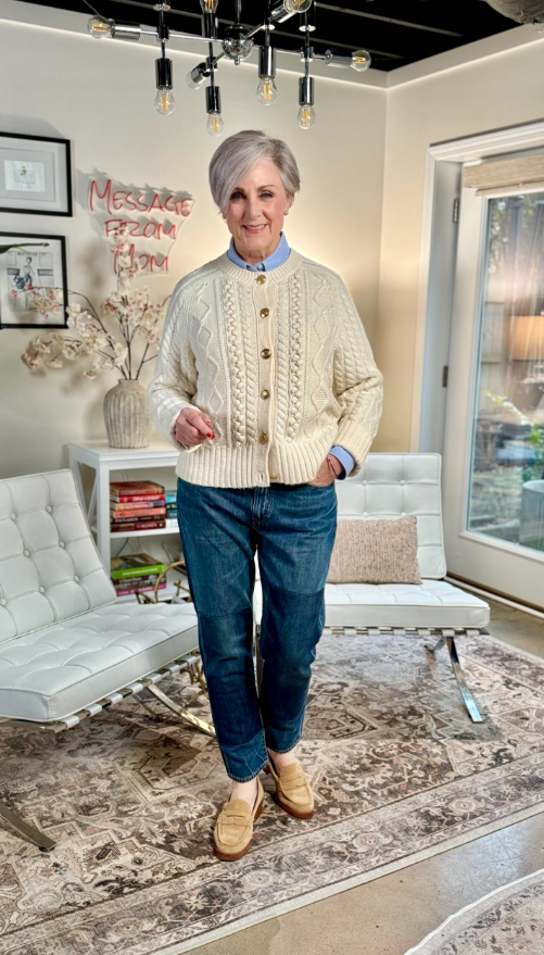 Woman wearing a cream cable-knit cardigan with gold buttons, cuffed blue jeans, and tan leather loafers, standing in a modern living space with neutral decor.