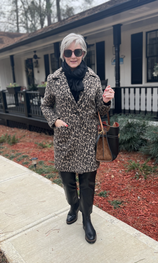 Woman wearing a leopard-print coat with a black scarf, black pants, and black knee-high boots, carrying a structured leather tote and walking along a sidewalk in front of a cottage-style home.