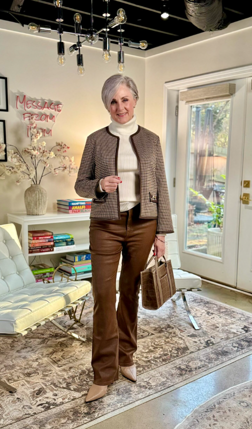 Woman indoors wearing a brown tweed jacket layered over a cream turtleneck, styled with brown leather pants, pointed-toe tan ankle boots with a low heel, and a structured brown handbag.