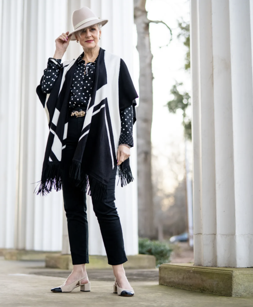 Woman wearing a black-and-white geometric wrap with fringe layered over a black polka-dot blouse and black pants, styled with a light felt fedora and block-heel pumps, standing between white columns.