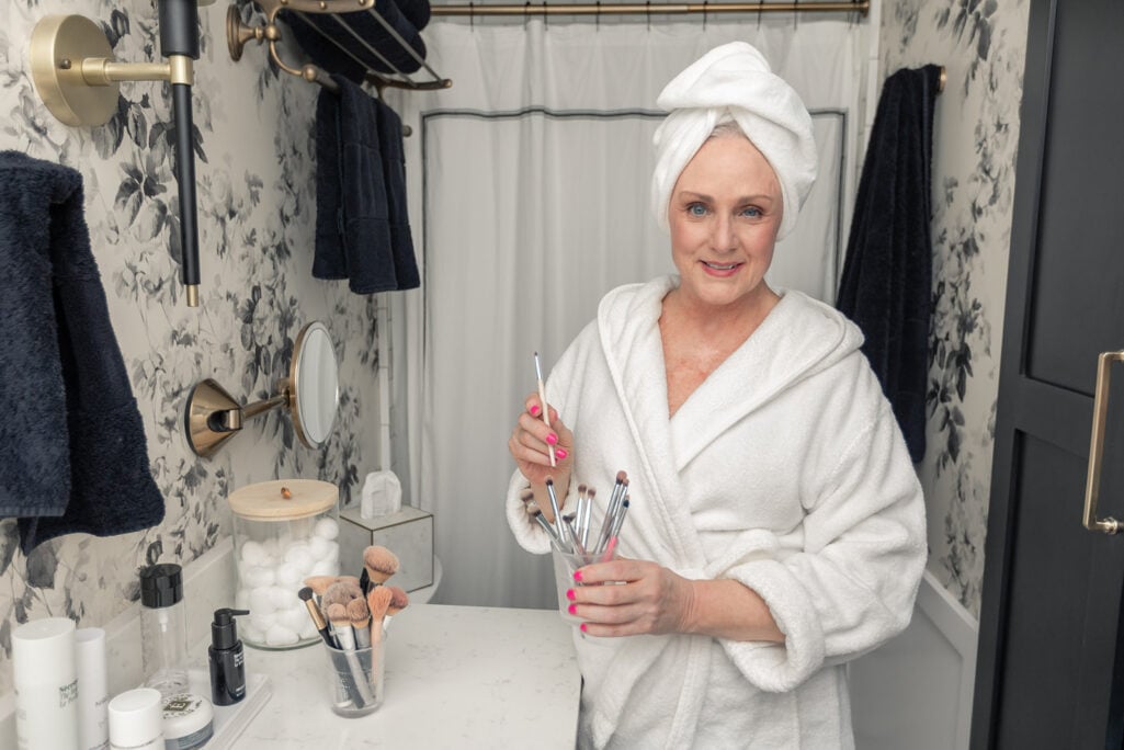 Woman over 50 in white bathrobe and hair towel holding makeup brushes in bathroom during skincare routine