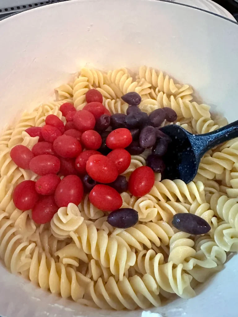Adding grape tomatoes and Kalamata olives to cooked fusilli pasta in a white pot