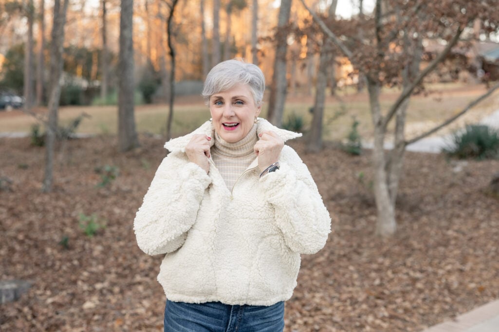 Woman over 50 in cream ribbed turtleneck layered under cream teddy fleece pullover with blue jeans