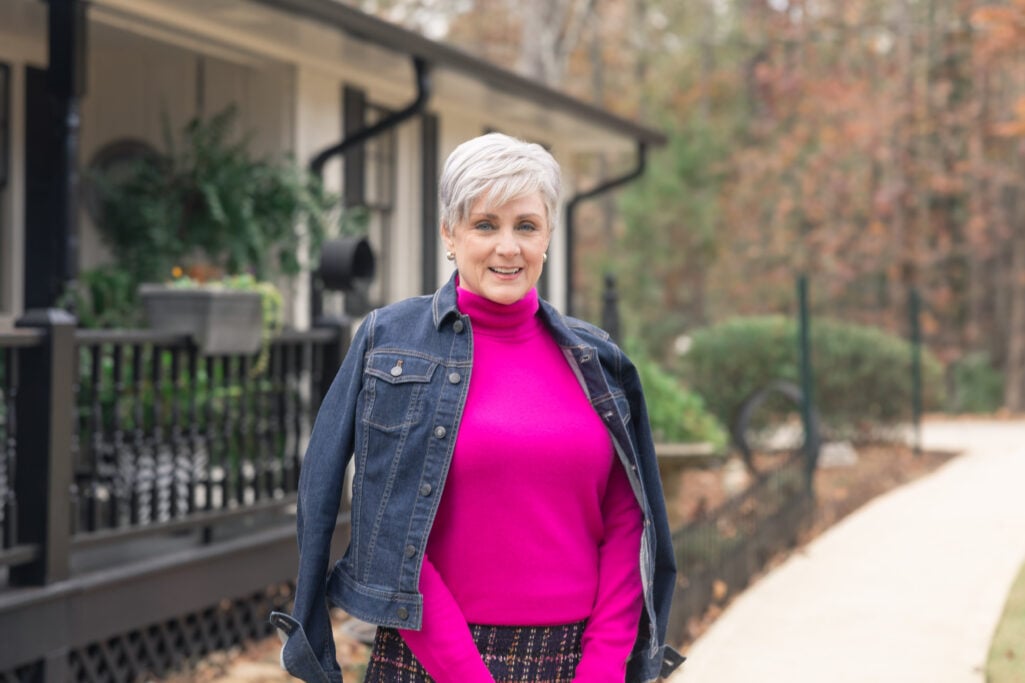 Woman over 50 wearing bright pink mock neck turtleneck under denim jacket with patterned tweed skirt