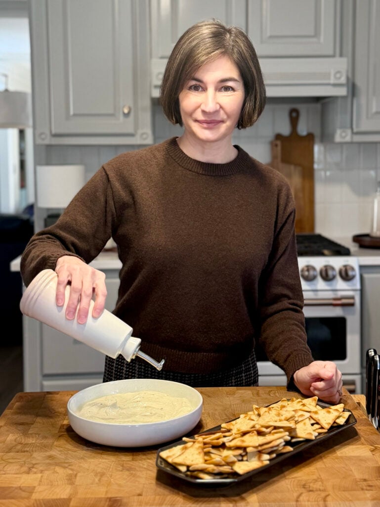 Kelly drizzling olive oil over whipped feta in the kitchen, with pita chips arranged on a black serving tray