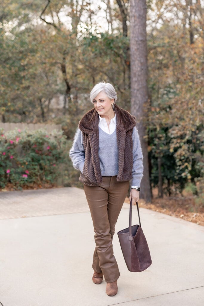 Woman over 50 styling white button-up under light blue V-neck sweater with fur vest, coated jeans, and suede ankle boots