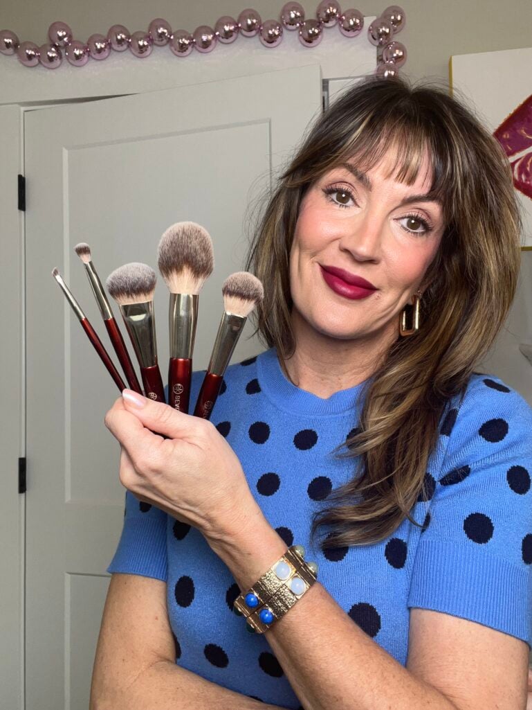 Woman holding bouquet of BK Beauty makeup brushes with burgundy handles, wearing blue polka dot sweater, berry lipstick, gold geometric earrings, and colorful gemstone bracelet
