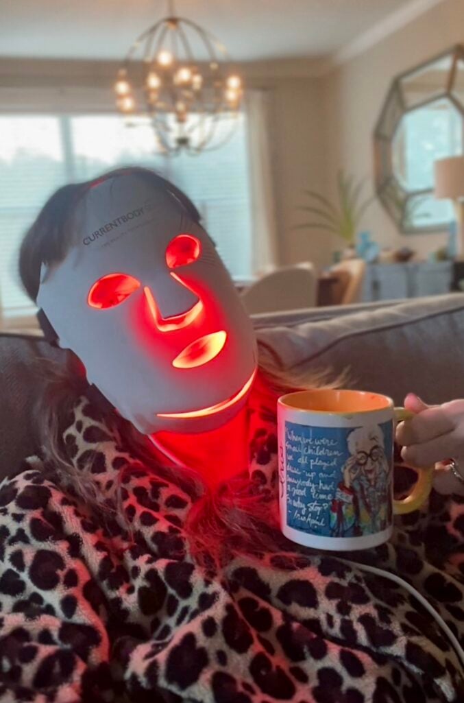 Woman using Currentbody red light LED face mask while relaxing on couch, holding illustrated coffee mug, wearing leopard print pajamas, with living room and chandelier visible in background