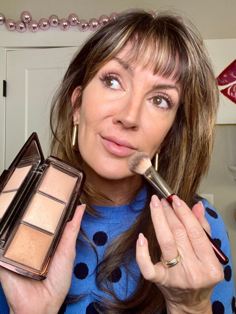 Woman holding open Hourglass Ambient Lighting powder trio palette in one hand and makeup brush against cheek in other, wearing blue polka dot sweater with natural glowing skin and soft pink lips