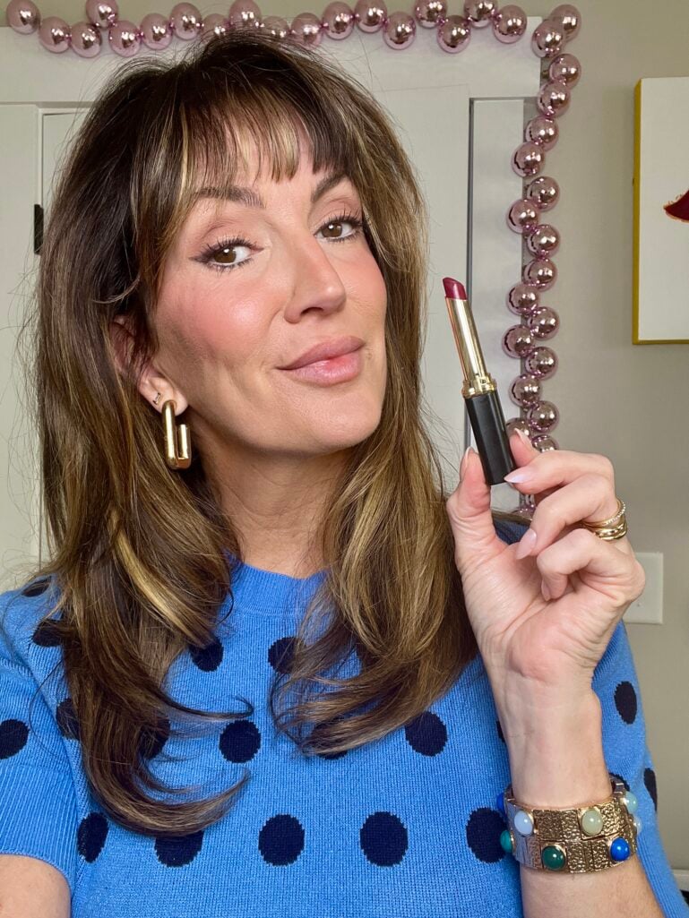 Woman holding L'Oreal berry lipstick up to camera, wearing blue polka dot sweater, gold geometric hoop earrings, stackable rings, and gemstone cuff bracelet, with lips bare showing before color