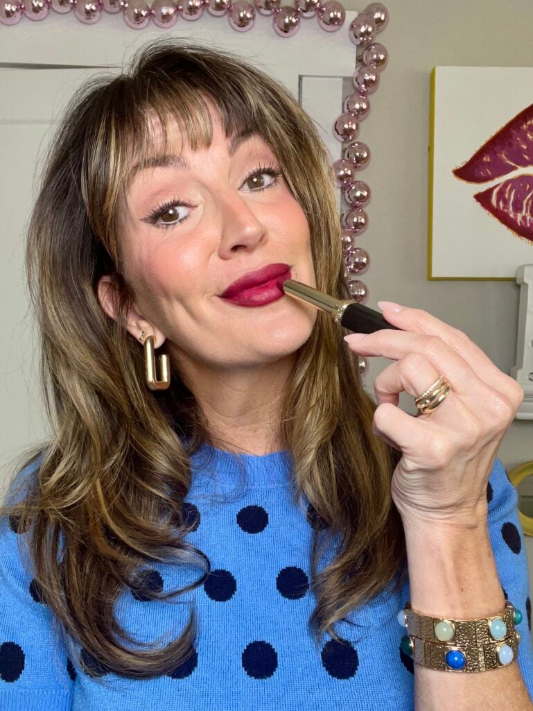 Woman applying berry matte lipstick while looking slightly off camera, wearing blue polka dot sweater, gold geometric earrings, and colorful beaded bracelet, with pink mirror frame visible behind