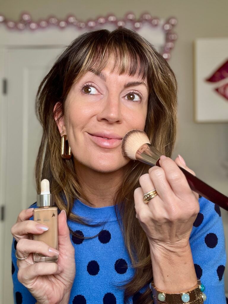 Woman holding foundation bottle with dropper and BK Beauty foundation brush against cheek, wearing blue polka dot sweater, gold geometric earrings, and leather cuff bracelet with natural makeup look