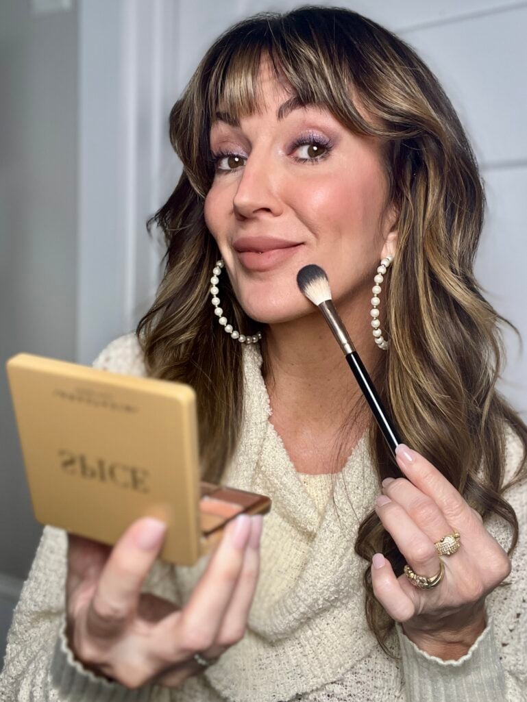 Woman with highlighted brown hair wearing cream knit sweater applying contour with black and white brush while holding open Anastasia Beverly Hills Spice bronzer palette in gold packaging, wearing purple shimmer eyeshadow, rosy cheeks, pearl hoop earrings, and gold stacked rings
