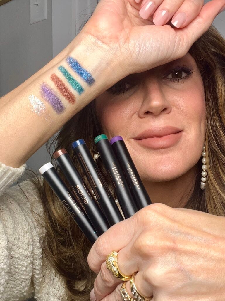 Woman displaying arm swatches of six Anastasia Beverly Hills Glidr shadow stick shades including white shimmer, purple, bronze, teal, and blue duo chrome while holding the corresponding sticks, wearing cream sweater, pearl hoop earrings, and gold rings