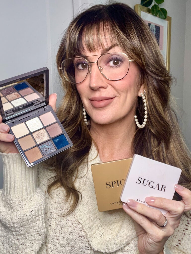 Woman wearing gold aviator glasses holding open Anastasia Beverly Hills Haze mini eyeshadow palette showing nine shades of neutrals and navy, plus closed Spice and Sugar mini palettes, wearing cream knit sweater, pearl hoop earrings, and purple shimmer eyeshadow