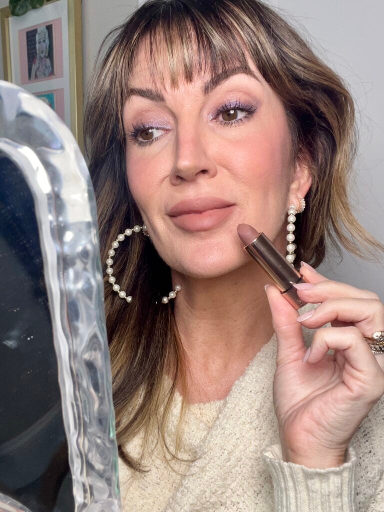 Woman holding Anastasia Beverly Hills matte lipstick bullet in nude-mauve shade Mauve Cloud near lips while looking in wavy glass mirror, wearing cream sweater, pearl hoop earrings, and purple shimmer eyeshadow, gallery wall visible in background