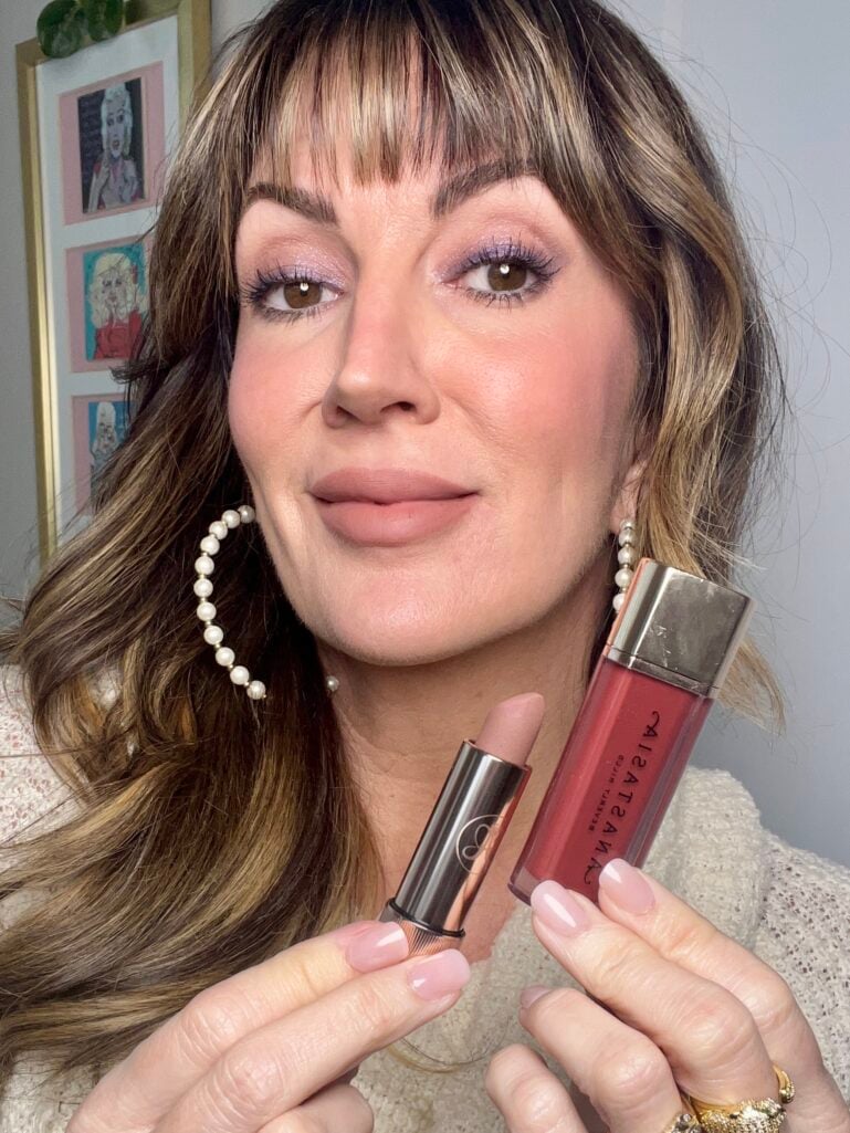 Woman holding Anastasia Beverly Hills nude lipstick bullet and berry-red Lip Velvet in shade Pomegranate, showing finished makeup look with purple shimmer eyes, rosy cheeks, and nude lips, wearing cream knit sweater and pearl hoop earrings, pop art gallery wall in background