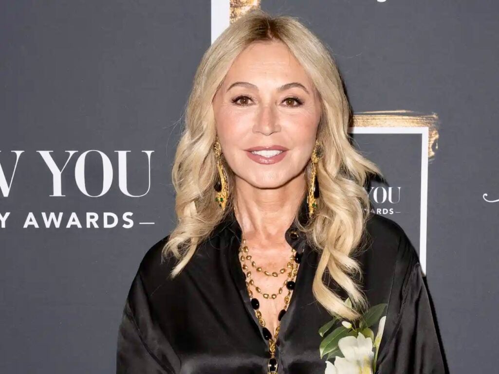 Anastasia Soare, founder of Anastasia Beverly Hills, at awards event wearing black silk blouse with gold and black beaded layered necklaces and matching drop earrings, blonde wavy hair, elegant neutral makeup