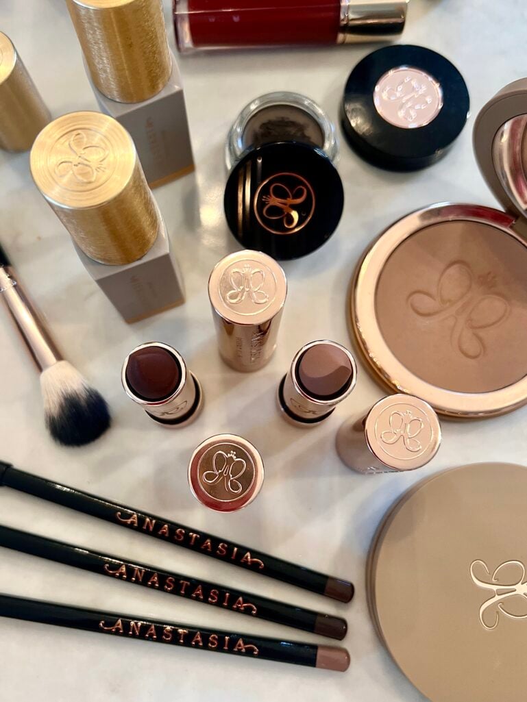 Close-up flatlay of Anastasia Beverly Hills products including Hydra Prime SPF 50, bronzer compact, Glidr shadow sticks in various shades, dipbrow pomade, lip products, and brow pencils on marble surface