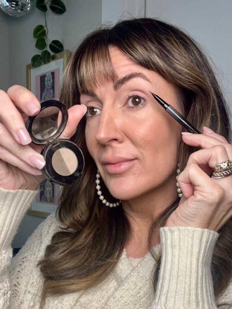 Woman holding Anastasia Beverly Hills brow powder duo compact in one hand and Micro Stroke brow pen near brow in other, demonstrating brow application technique, wearing cream knit sweater, pearl hoop earrings, and gold rings, with gallery wall and trailing plant visible in background