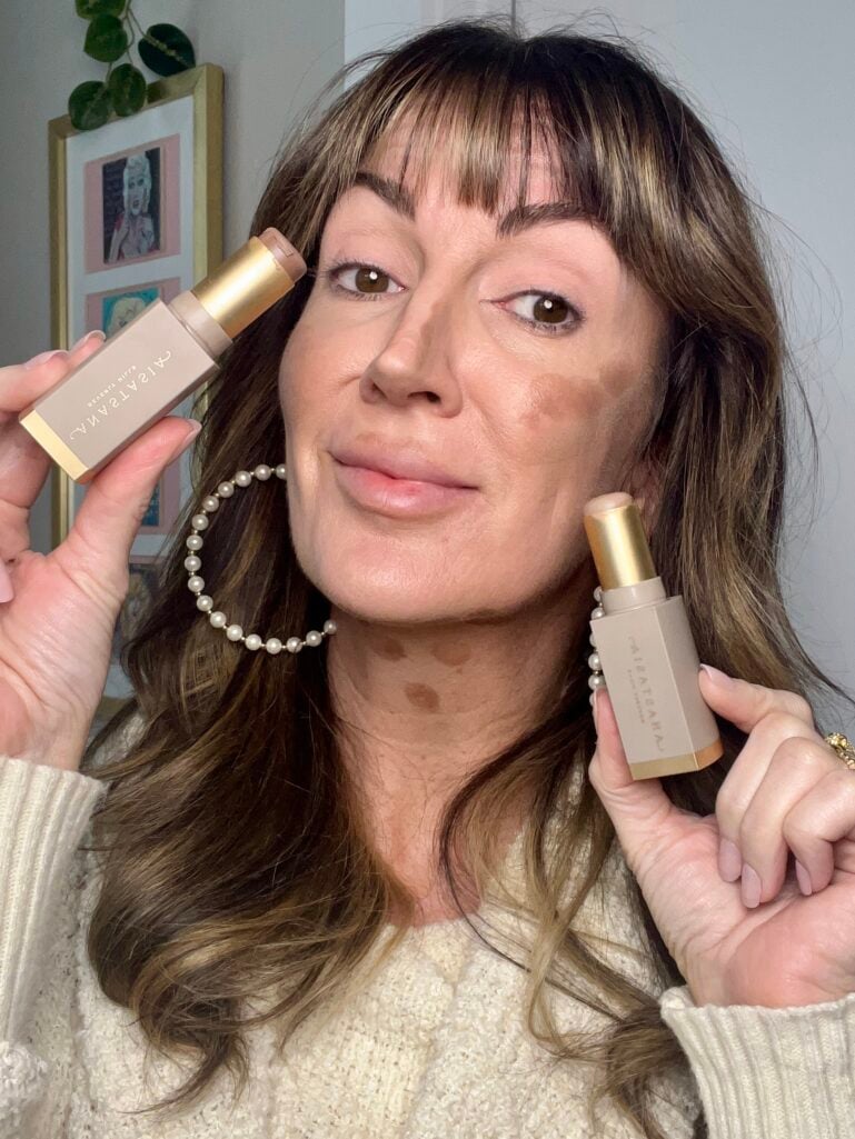 Woman holding two Anastasia Beverly Hills Smooth Blur contour sticks near face showing unblended contour product applied to cheeks and forehead, wearing cream knit sweater and pearl hoop earrings, demonstrating contour placement before blending
