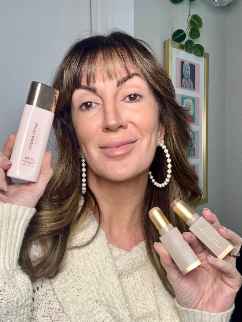 Woman holding Anastasia Beverly Hills Hydra Prime SPF 50 bottle in one hand and two contour sticks in other, showing natural radiant skin with no foundation, wearing cream knit sweater and pearl hoop earrings, gallery wall with pop art portraits in background