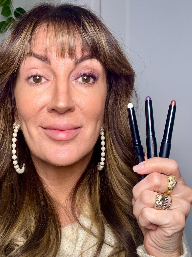 Woman holding three Anastasia Beverly Hills Glidr shadow sticks showing purple, bronze, and duo chrome blue shades, wearing purple shimmer eyeshadow look, cream knit sweater, pearl hoop earrings, and gold stacked rings