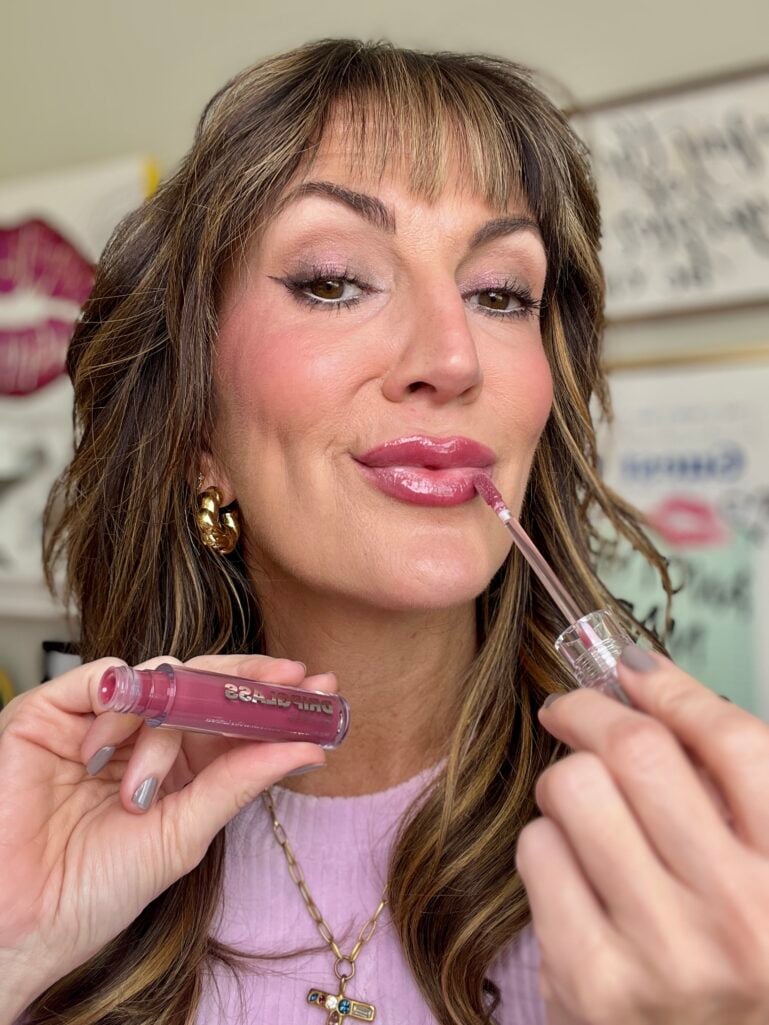 Woman applying Morphe Glazed Drip glass gloss in shatterproof mauve shade with doe-foot applicator, wearing lilac sweater with gold twisted earrings, showing glossy pink lip result with shimmer eyeshadow look