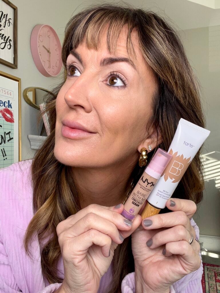 Woman looking to the side holding NYX Bare With Me concealer serum and Tarte BB Blur tinted moisturizer near face, wearing lilac sweater with gold knot earrings, gallery wall with pink clock and typography prints visible in background