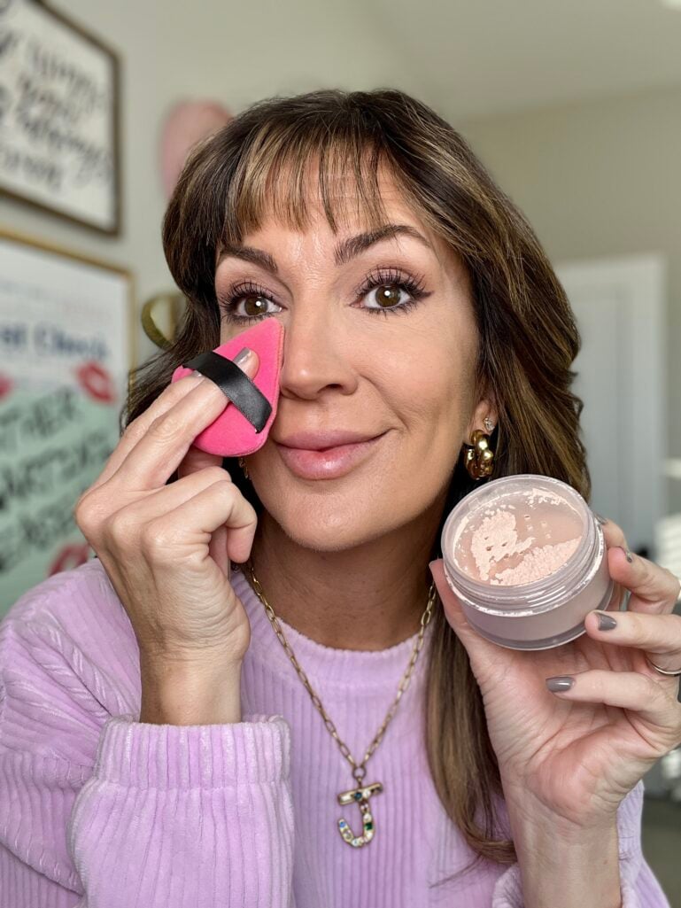 Woman pressing hot pink triangle powder puff to under-eye area while holding open jar of Laura Mercier Ultra Blur translucent loose setting powder in rose shade, wearing lilac sweater and jeweled J initial necklace, demonstrating powder application technique