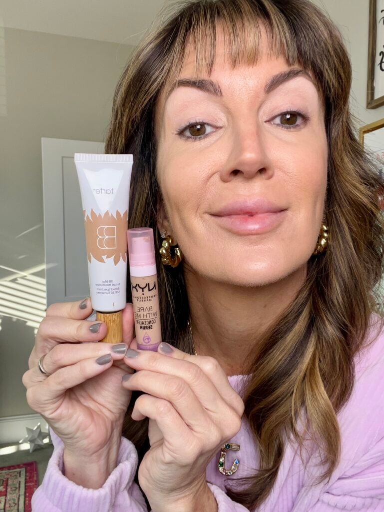 Woman holding Tarte BB Blur tinted moisturizer and NYX Bare With Me concealer serum up to camera, showing natural bare-faced skin prep, wearing lilac sweater, gold knot earrings, and jeweled J initial necklace