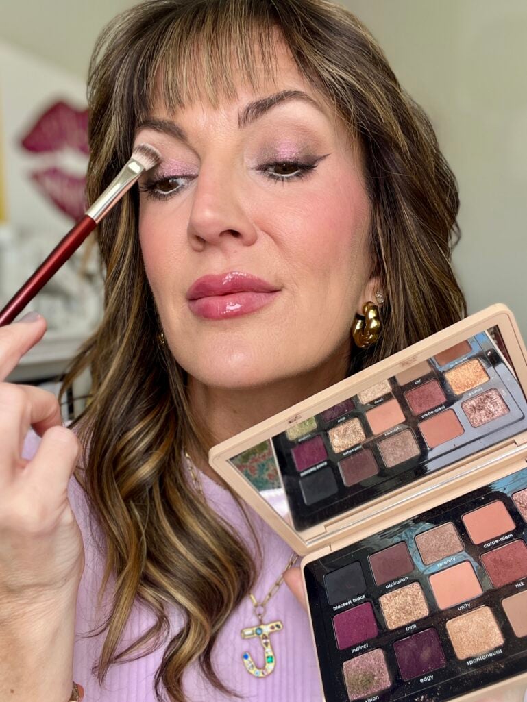 Woman applying eyeshadow with burgundy-handled brush while holding two Natasha Denona palettes including My Dream Palette and mini eye sculpt palette, showing purple-pink shimmer eyeshadow look, wearing lilac sweater with glossy pink lips