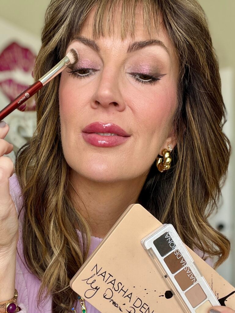 Close-up of woman blending eyeshadow on lid with BK Beauty brush while holding Natasha Denona My Dream Palette and mini sculpt palette, showing soft purple-mauve shimmer eye look, wearing gold twisted hoop earrings and glossy lips