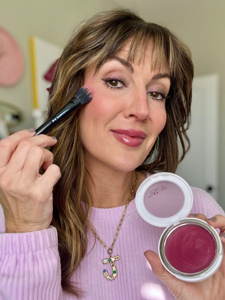 Woman applying cream blush to cheek with e.l.f. putty blush brush while holding open Makeup by Mario Soft Pop Plumping Blush Veil compact in berry punch shade, wearing lilac sweater with jeweled J initial necklace, showing rosy flush result