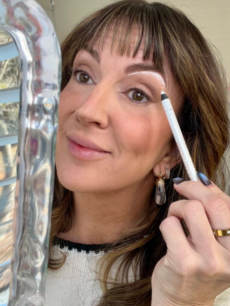 Woman applying white Makeup Revolution eyeliner pencil under brow bone while looking in iridescent handheld mirror, showing brow lift technique, wearing cream sweater with black trim and blue-gray press-on nails