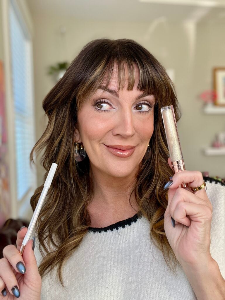 Woman smiling holding white eyeliner pencil and gold shimmer lip gloss, displaying finished Pantone Cloud Dancer inspired makeup look with glossy lips and luminous skin