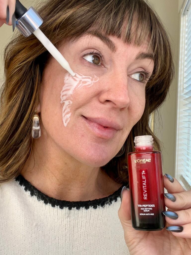 Woman applying milky white L'Oreal Paris Revitalift Triple Power Tri-Peptides serum to cheek with dropper, holding red serum bottle, demonstrating skincare application