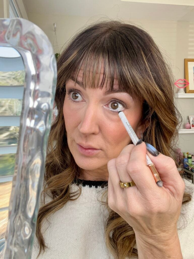 Woman applying white eyeliner to lower waterline while looking in mirror, demonstrating eye-brightening tightlining technique with Makeup Revolution pencil