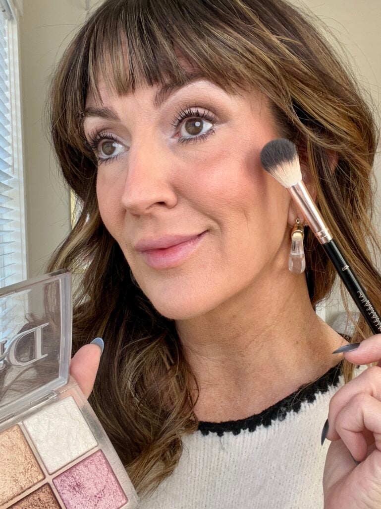 Woman applying highlighter to cheekbone with Anastasia Beverly Hills tapered powder brush while holding Dior Backstage Glow quad palette showing white, champagne, pink, and bronze shades