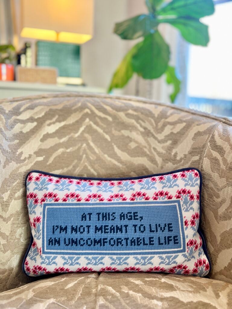 Furbish Studio needlepoint pillow reading "At This Age, I'm Not Meant To Live An Uncomfortable Life" in blue with pink and light blue floral cross-stitch border, displayed on beige zebra print upholstered chair with houseplant and lamp in background