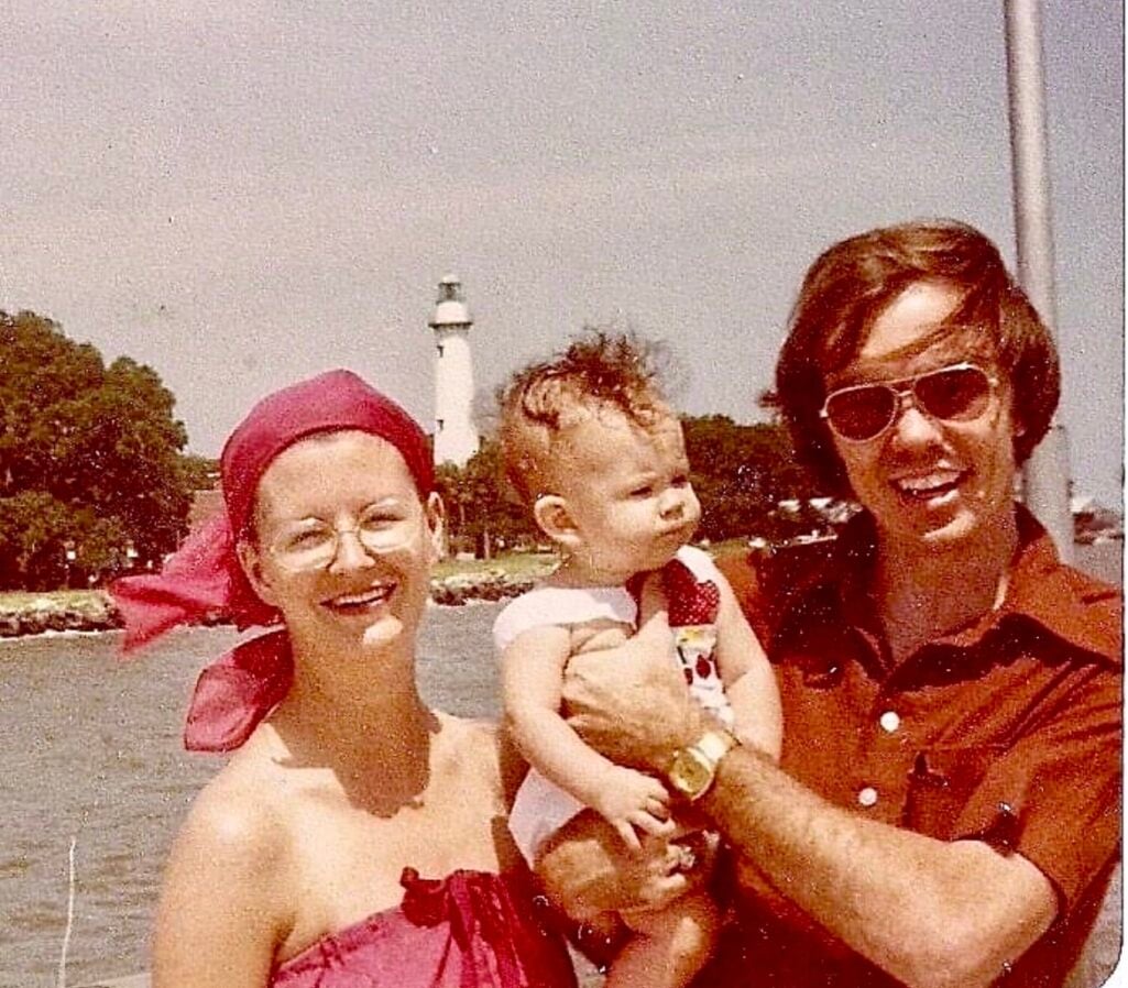 Vintage 1970s family photo of baby Jennifer being held by her parents near a lighthouse, mother wearing magenta swimsuit and matching head scarf with oversized glasses, father in rust-colored polo shirt and aviator sunglasses with gold watch