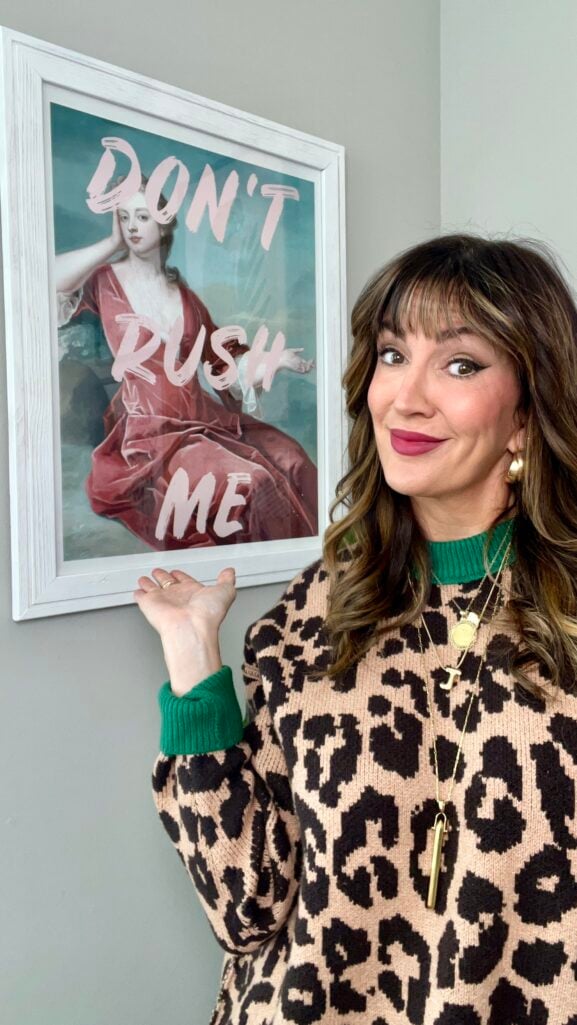 Woman standing next to framed art print reading "Don't Rush Me" featuring classical portrait with pink text overlay, wearing leopard print sweater with green ribbed trim, gold hoop earrings, layered gold necklaces including J initial pendant, and berry lipstick