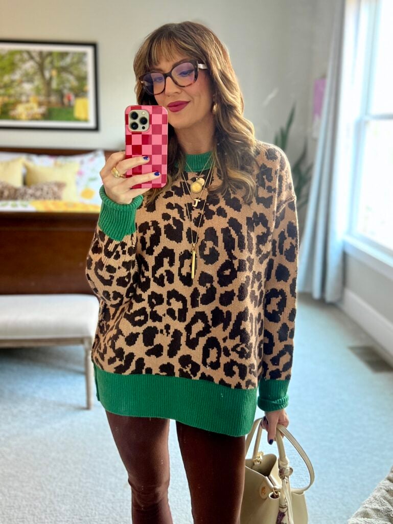 Mirror selfie of woman wearing oversized Aerie leopard print sweater with green ribbed collar, cuffs, and hem, burgundy leather pants, tortoise cat-eye glasses, layered gold necklaces, holding beige mini bucket handbag with scarf detail, pink checkered phone case visible