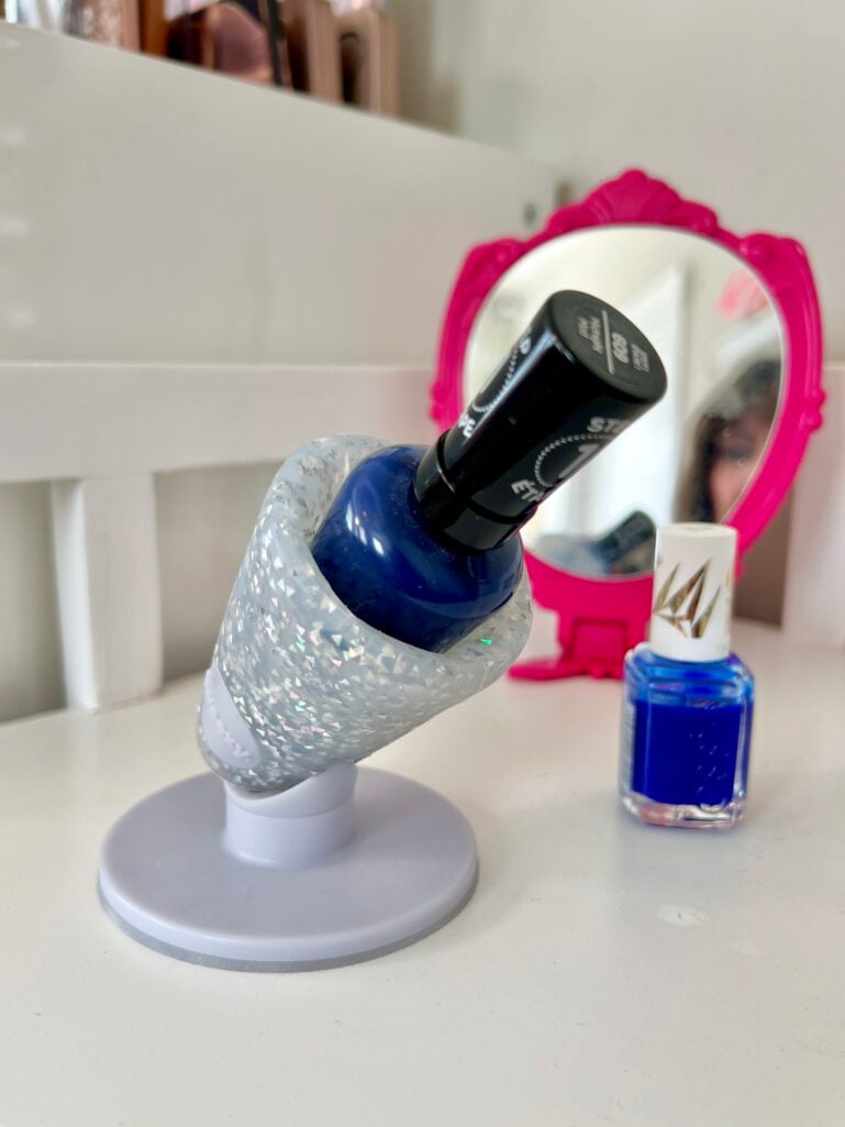 Untippable nail polish holder in white glitter finish gripping dark blue Essie polish bottle at an angle on vanity surface, with royal blue Essie polish bottle beside it and hot pink decorative mirror in background