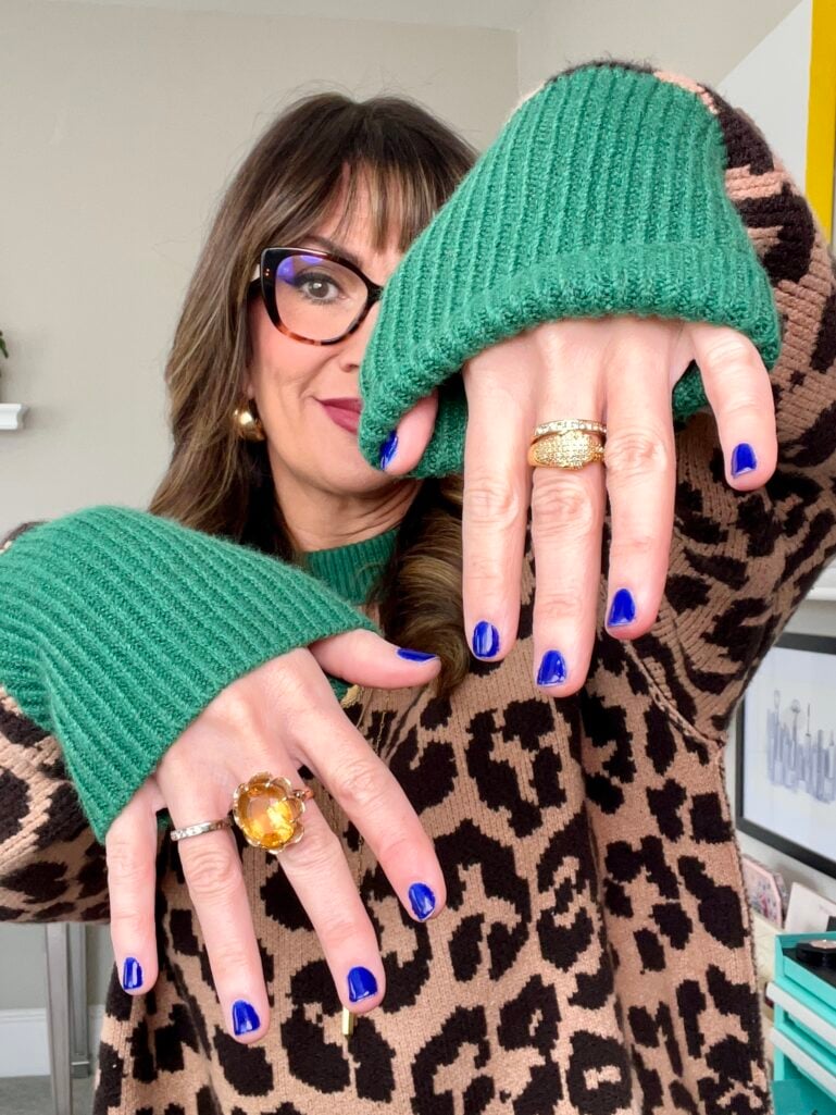 Woman displaying both hands with short nails painted in royal blue Essie polish, wearing leopard print sweater with green ribbed cuffs, tortoise glasses, gold stacked rings including large citrine flower cocktail ring, berry lipstick